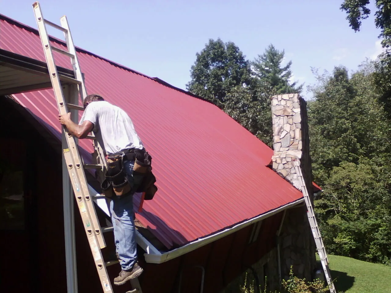 Roofer on a ladder beside a red metal roof and stone chimney — Soffit & Fascia Repair by Roofing Brothers in Cobb Island